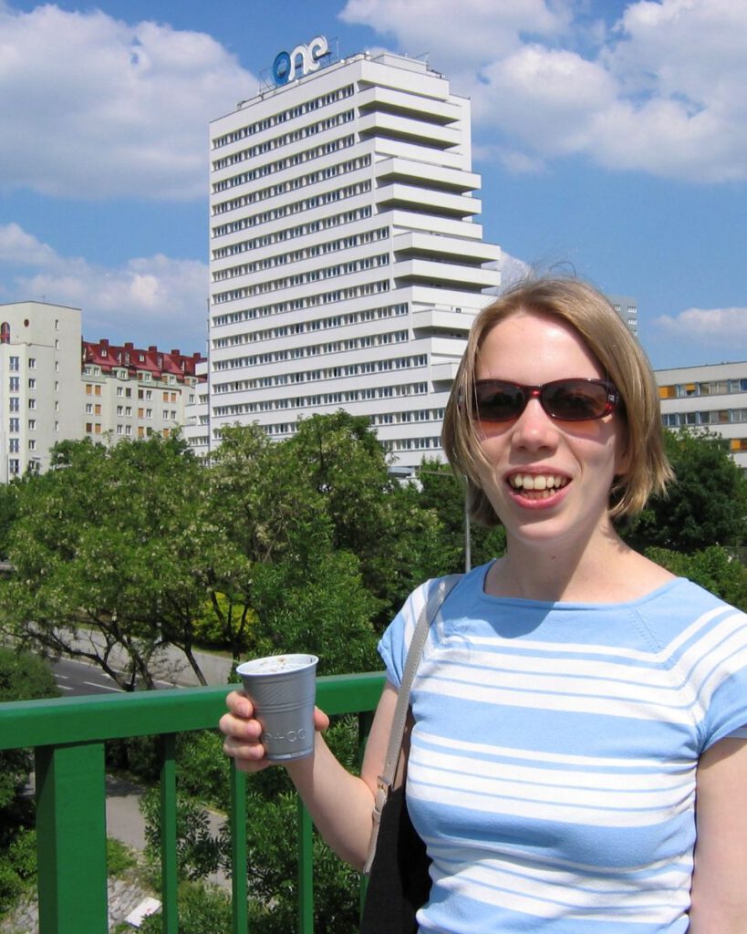 Astrid 2004 vor ihrem Studentenwohnheim in Wien, sie trägt eine Sonnenbrille und hat einen To-Go-Becher Kaffee in der Hand