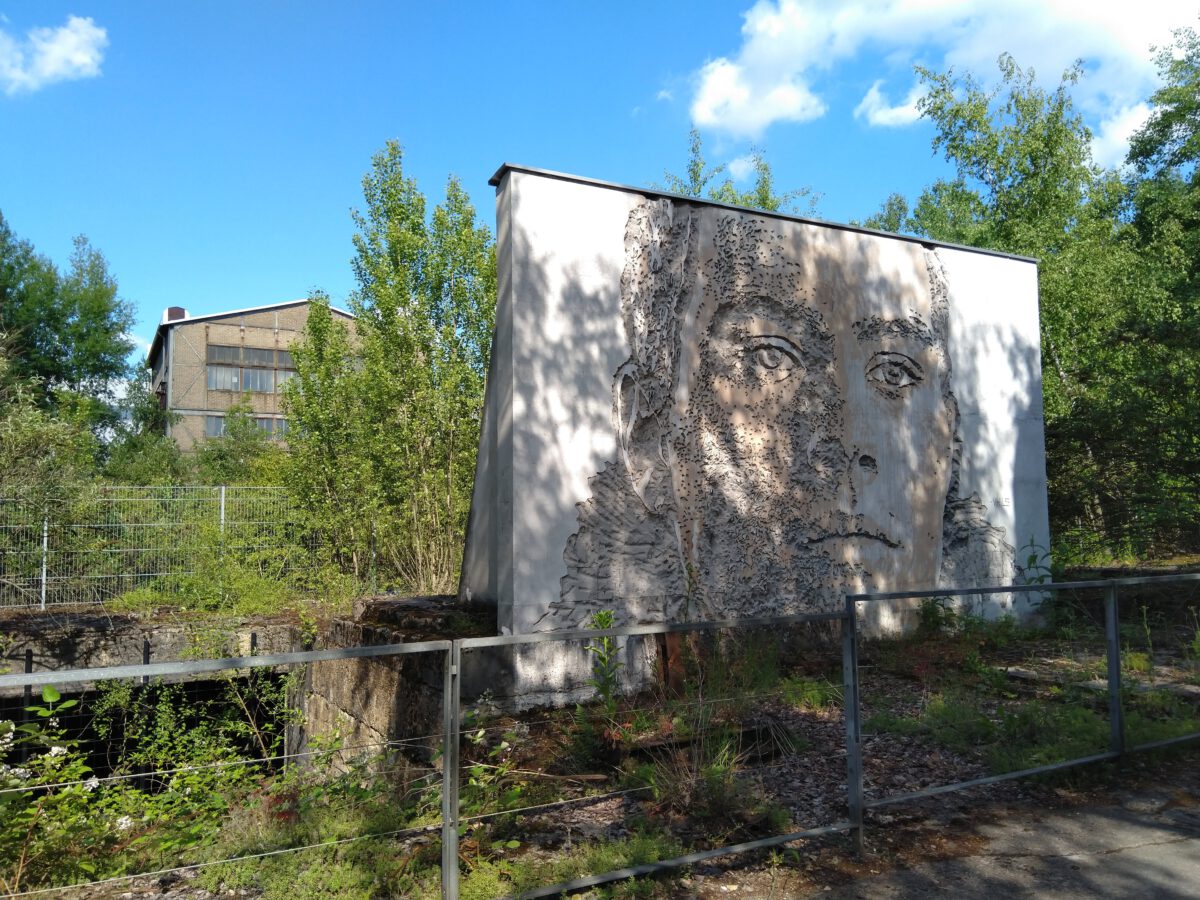 Foto von einem verwilderten Industriegelände. Im Vordergrund steht eine Betonwand, in die mit einem Meißel ein Gesicht geschlagen wurde.