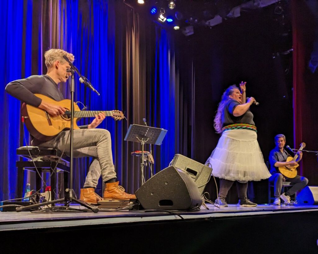 Anna Mateur mit ihren beiden Gitarristen auf der Bühne des Neuen Theater Höchst. Sie trägt einen Reifrock und bunte Hoola-Reifen.