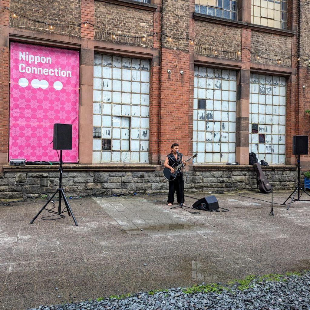 Ein Gitarrist mit Verstärkern vor der Naxoshalle im Hintergrund ein großes Nippon Connection Banner