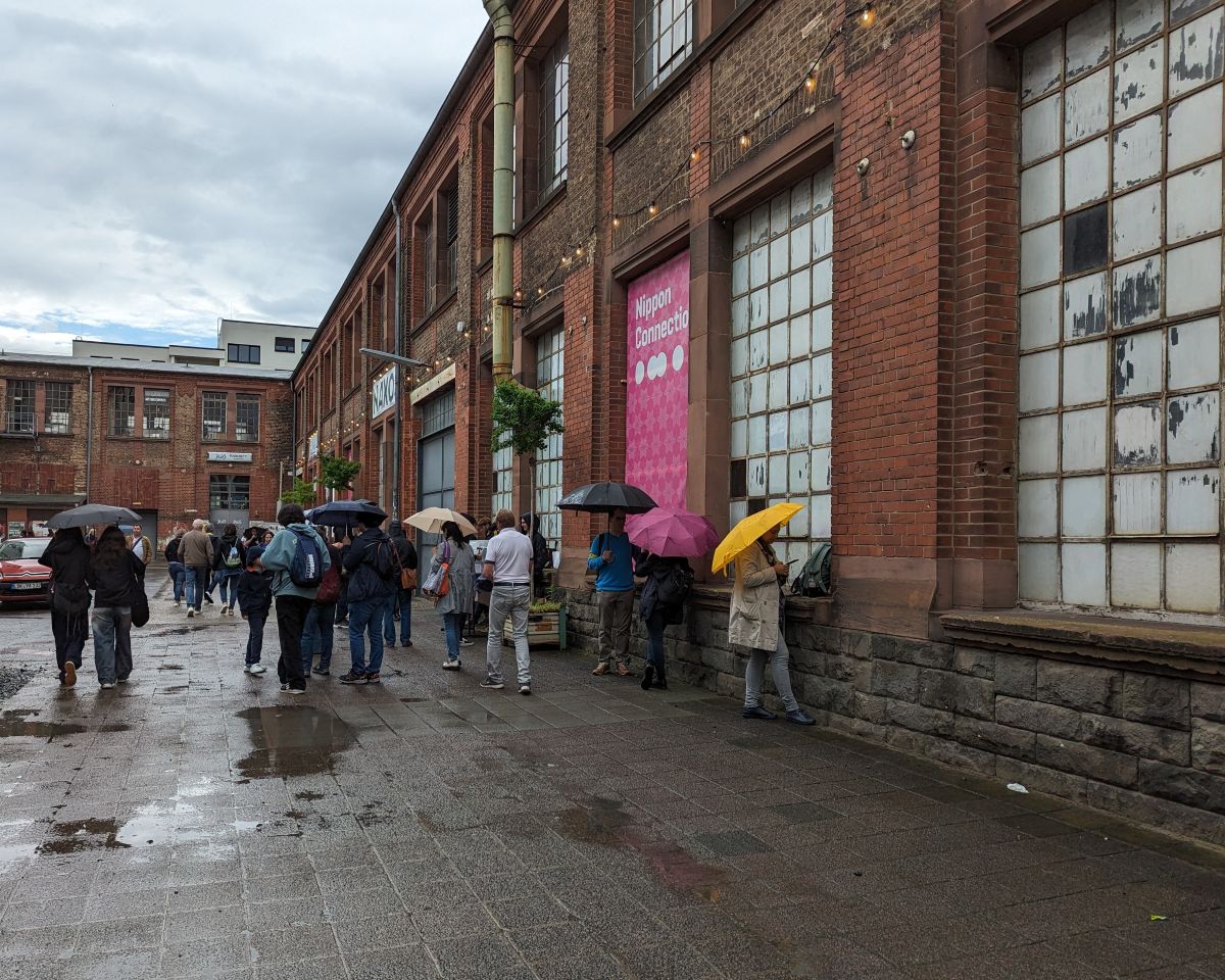 Das Naxos Kulturgelände bei Regen: Es haben sich vor der Fabrikhalle Pfützen gebildet und die Passanten haben ihre Regenschirme aufgespann.