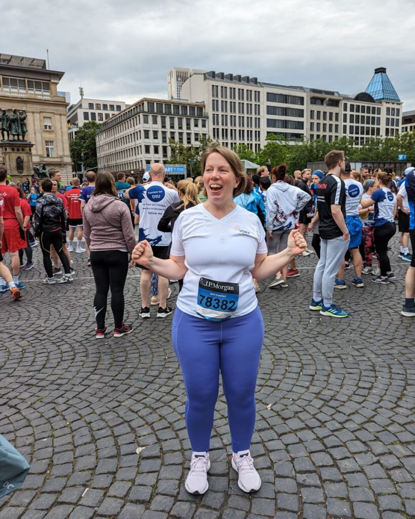 Astrid mit Firmen-T-Shirt, lila Leggins und Startnummer für den J. P. Morgan Lauf 2024 beim Warten auf den Start.