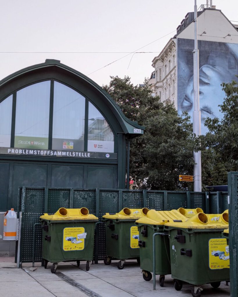 Bild eines kleinen Gebäudes auf dem Wiener Naschmarkt mit der Aufschrift "Problemstoffsammelstelle", davor stehen große Müllcontainer. Im Hintergrund ist ein großes Mural an einem Altbau zu erkennen: Ein Gesicht kopfunter mit geschlossenen Augen.