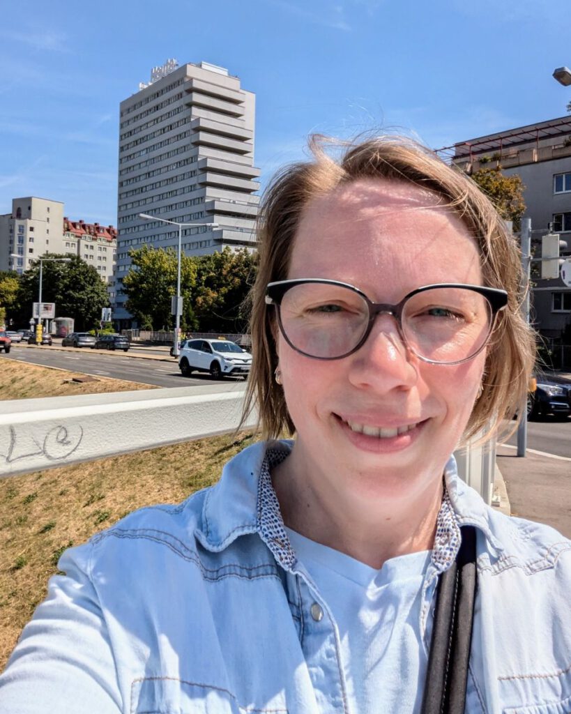 Astrid vor ihrem alten Studentenwohnheim in Wien, 2024. Es ist ein Selfie und man erkennt, dass es ein sehr heißer Tag ist.