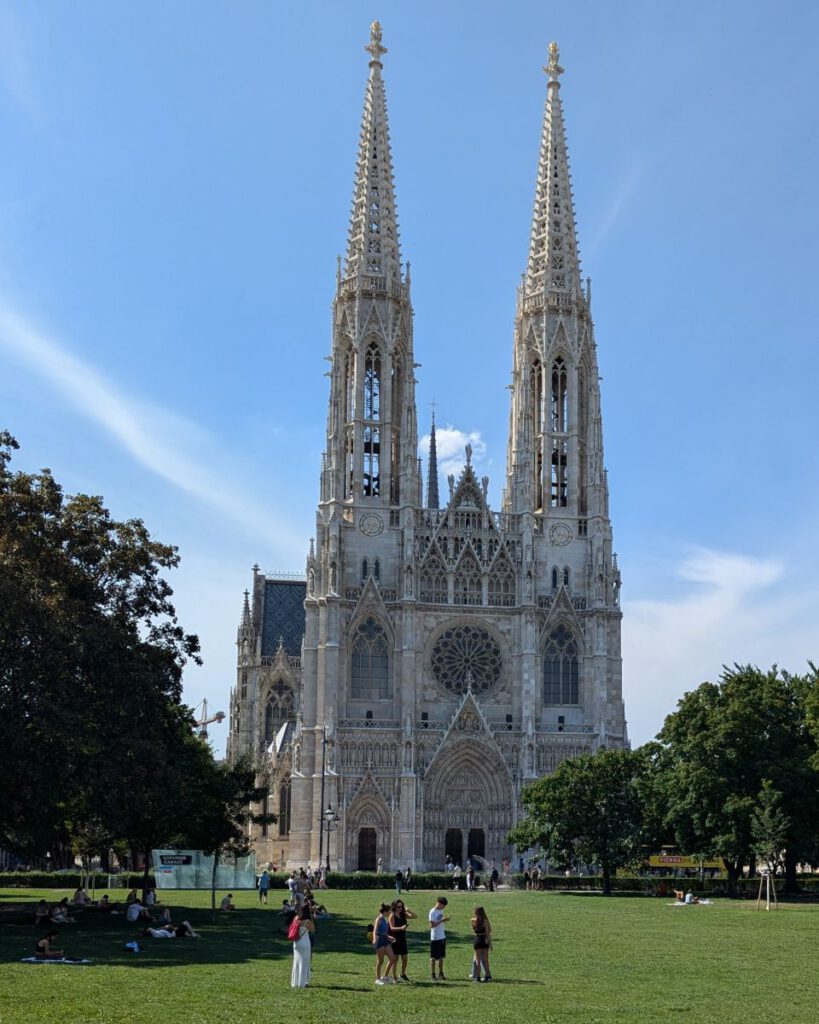 Foto der Votivkirche in Wien, davor eine belebte Rasenfläche mit schattigen Bäumen.