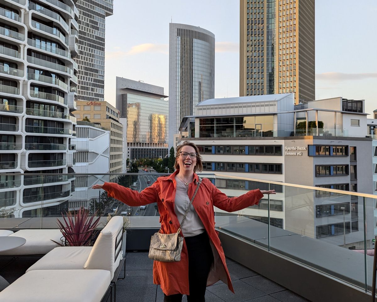 Astrid auf einer Dachterasse in Frankfurt. Sie grinst, trägt einen orangefarbenen Trechcoat und in der Skyline hinter ihr spiegelt sich die Sonne.