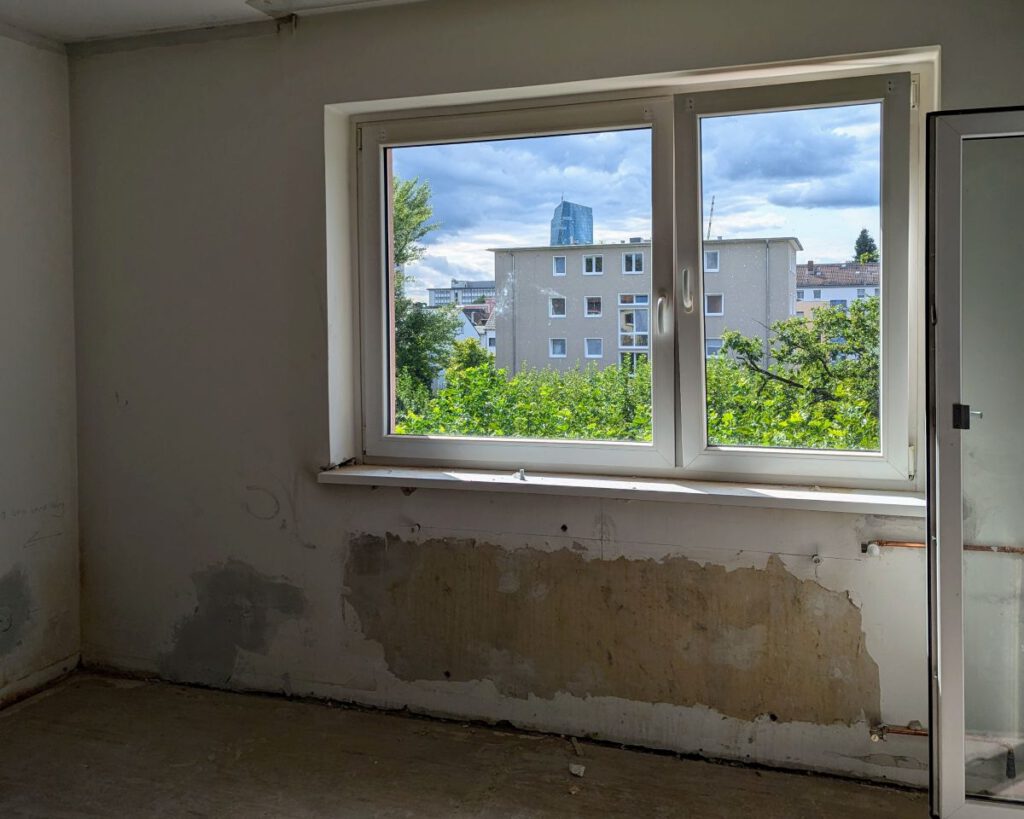 Blick aus einem dunklen Raum mit abgerissener Tapete, ohne Bodenbelag und mit nackten Heizungsrohren. Aus dem Fenster sieht man Bäume und in der Ferne die Europäische Zentralbank.