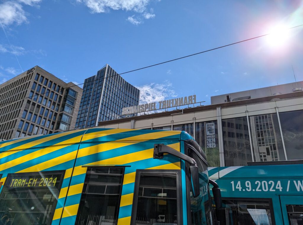 Bild von zwei Straßenbahnen vor dem Frankfurter Schauspielhaus. Auf der einen Tram steht Tram-EM 2024 auf der anderen sieht man das Datum 14.9.2024. Der Himmel ist blau, die Sonne scheint.