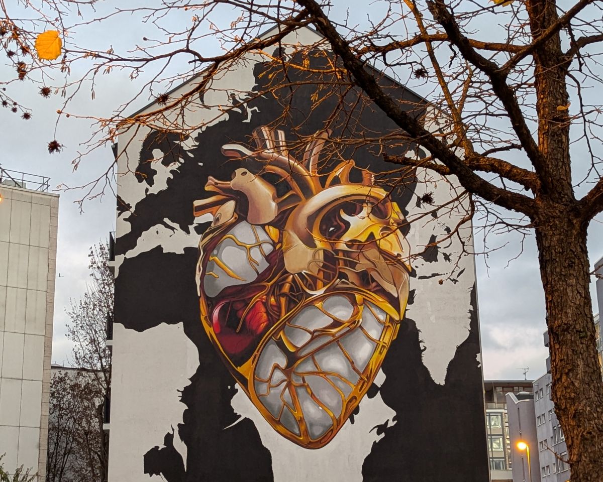 Mural in Frankfurt, in der Dämmerung aufgenommen. Man erkennt ein großes stilisiertes Herz in Goldfarben. Davor ein kahler Baum, an dem nur noch wenige gelbe Blätter hängen.