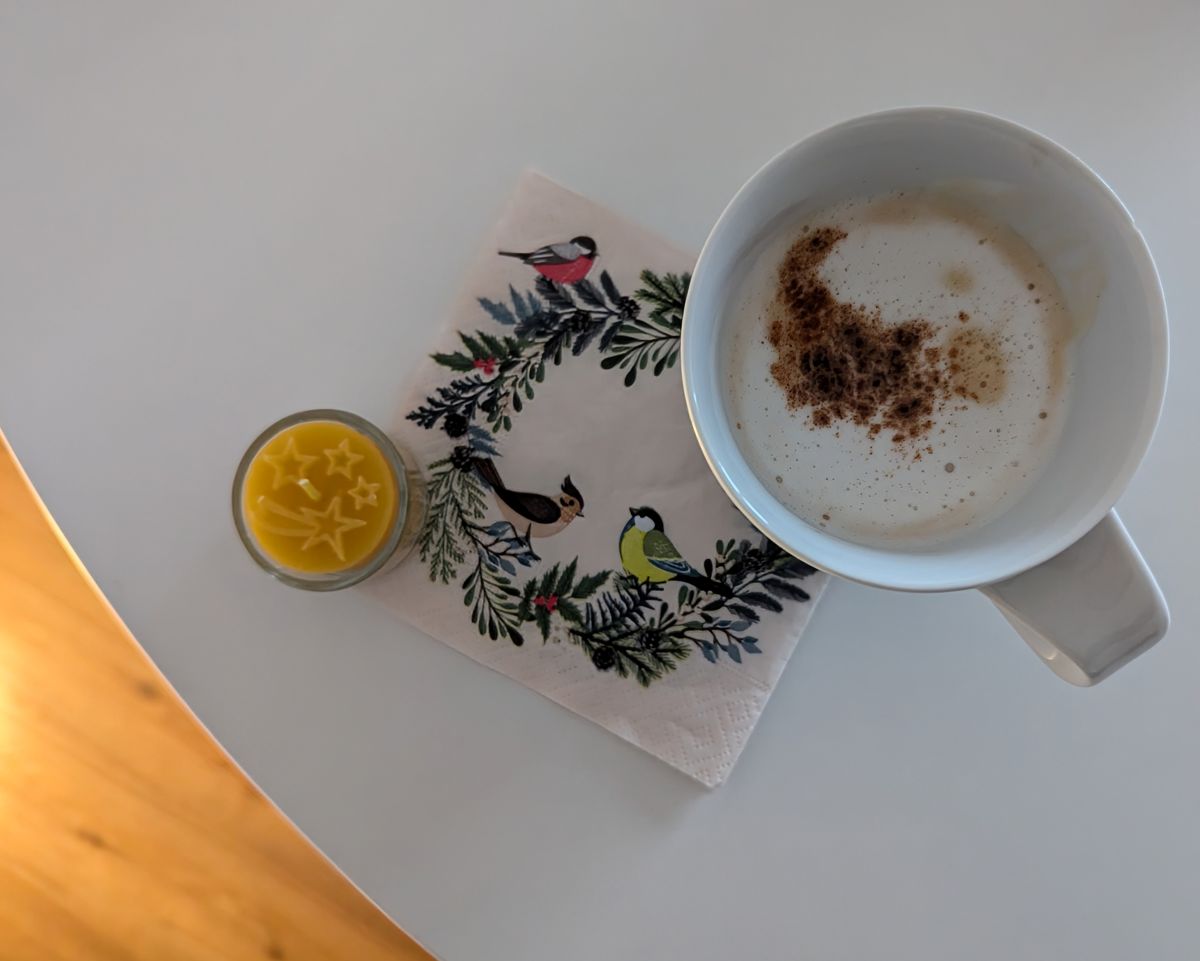 Runder Tisch, auf dem eine Kaffeetasse mit Milchschaum und Zimt steht, darunter eine kleine Serviette mit einem kleinen Kranz bedruckt.