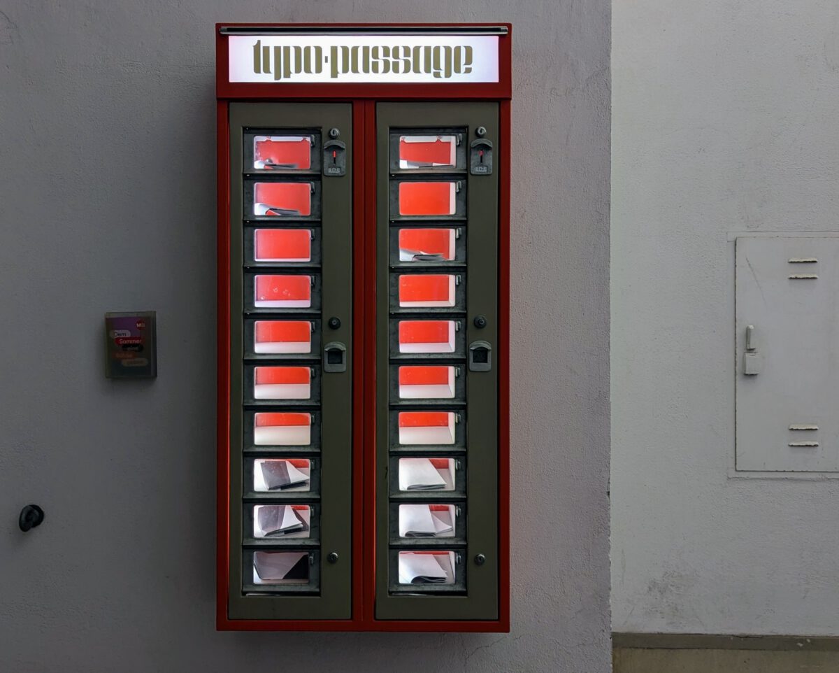 Das Bild zeigt einen klassischen roten Verkaufsautomaten aus den 60er Jahren. Er wird hier aber verwendet Kunst zu verkaufen. Am Automat steht "Typopassage"