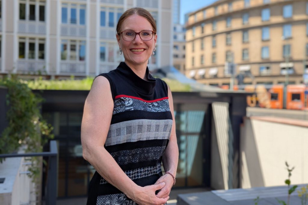 Portraitaufnahme von Maren Hünnerscheidt, im Hintergrund erkennt man die Dachbegrünung des Anbaus und eine Straßenhan. Mahren lächelt strahlend, ihre Schulter wird von der Sonne angeleuchtet.