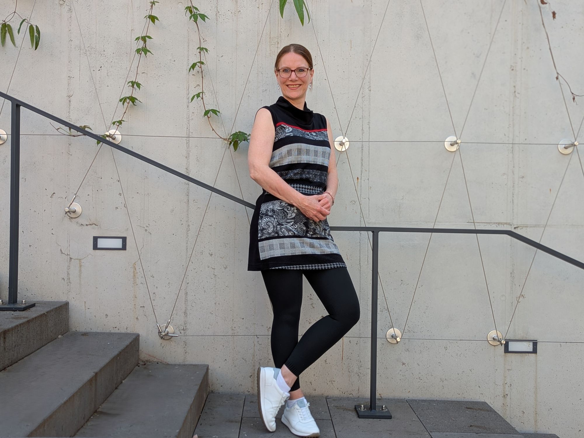 Maren Hünnerscheidt steht locker auf einer Treppe und blickt offen und lächelnd in die Kamera, ihre weißen Turnschuhe blitzen auf dem grauen BetonIm Hintergrund ranken zarte Blätter.