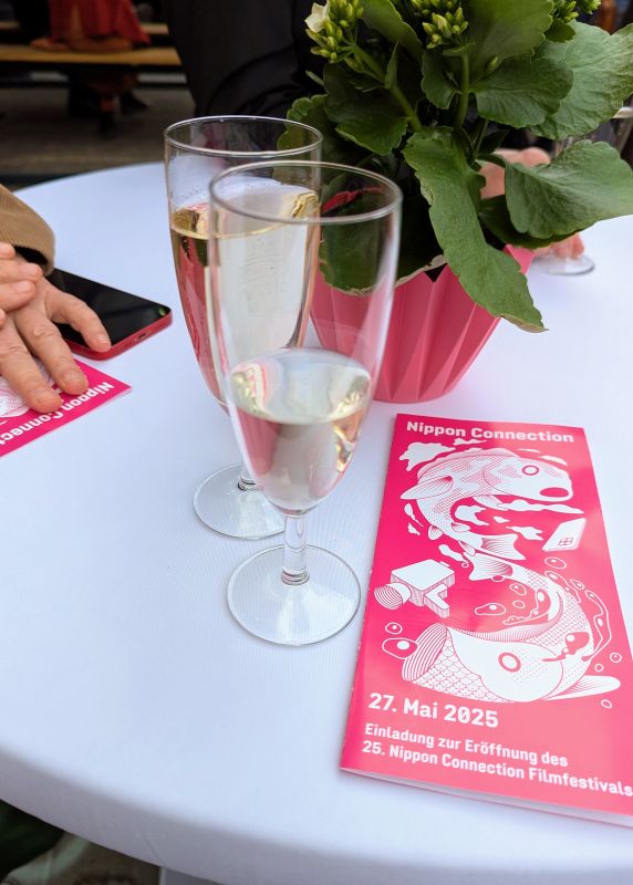 Empfang vor dem Mousonturm. Auf einem Stehtisch liegt die Einladung und es steht ein Glas Pflaumenwein und ein Glas Sake auf dem Tisch.