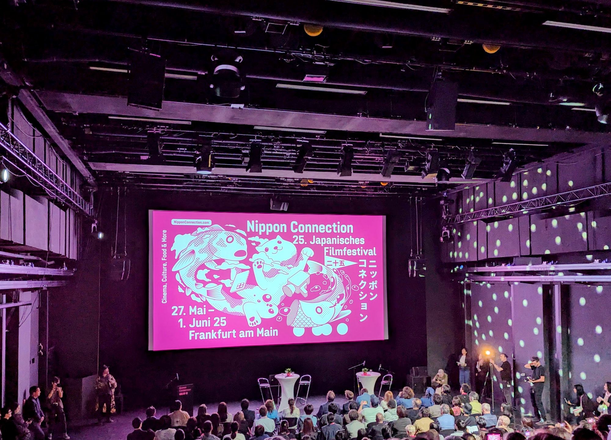 Leinwand im Mousonturmsaal. Es wird das pinke Design des Nippon Connectiion Festivals gezeigt. Davor eine Bühne mit zwei Stehtischen und einem Redepult