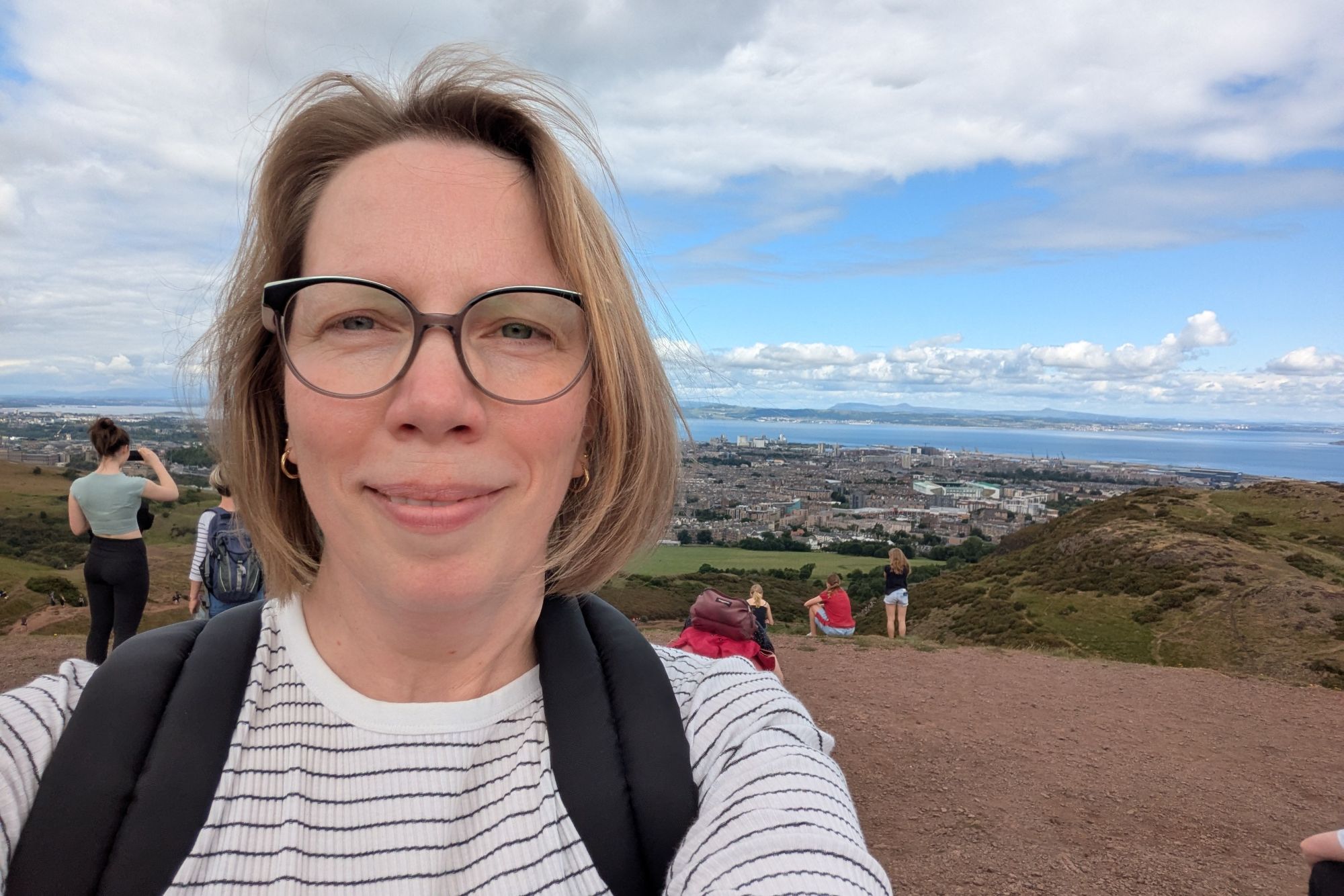 Astrid macht ein Selfie auf dem Gipfel von Arthurs Seat, im Hintergrund Wolken und das Meer.
