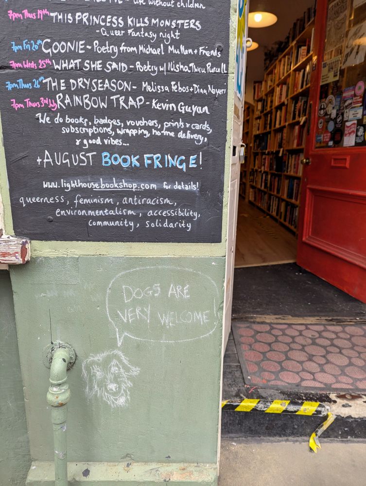 Eingang des Lighthouse Bookshops, an eine rTafel sind die Neuerscheinungen angeschrieben und darunter mit Kreide ein wuscheliger Hund, der sagt "Dogs are welcome here).