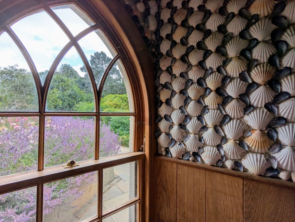 Die kleine Gartenhütte von innen, aus dem Fenster schaut man auf Lavendel-Rosenbeete, die Wände sind mit Muscheln verziert.