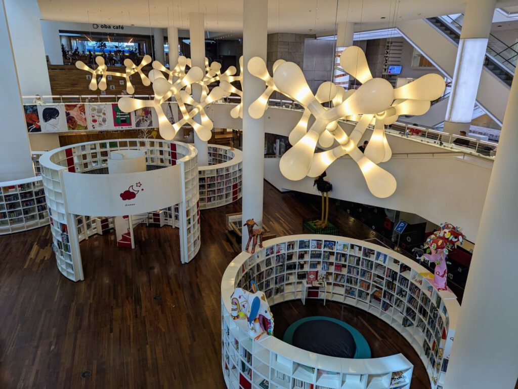 Blick von der Empore auf den Kinderbereich der Bibiliothek. Runde Regale mit Büchern, in deren Mitte kleine Leseinseln und oder eine Wendeltreppe nach oben ist.