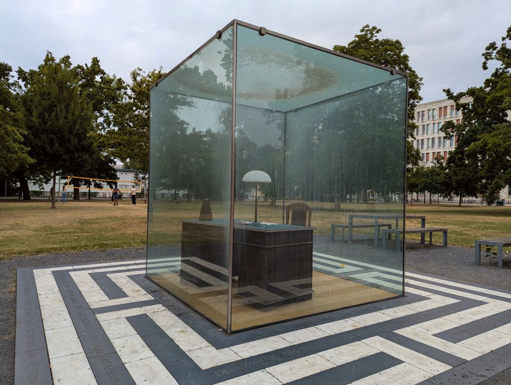 Das Adorno-Denkmal: Schreibtisch von Theodor W. Adorno in einem Glaskasten. Im Hintergrund wird Volleyball gespielt.