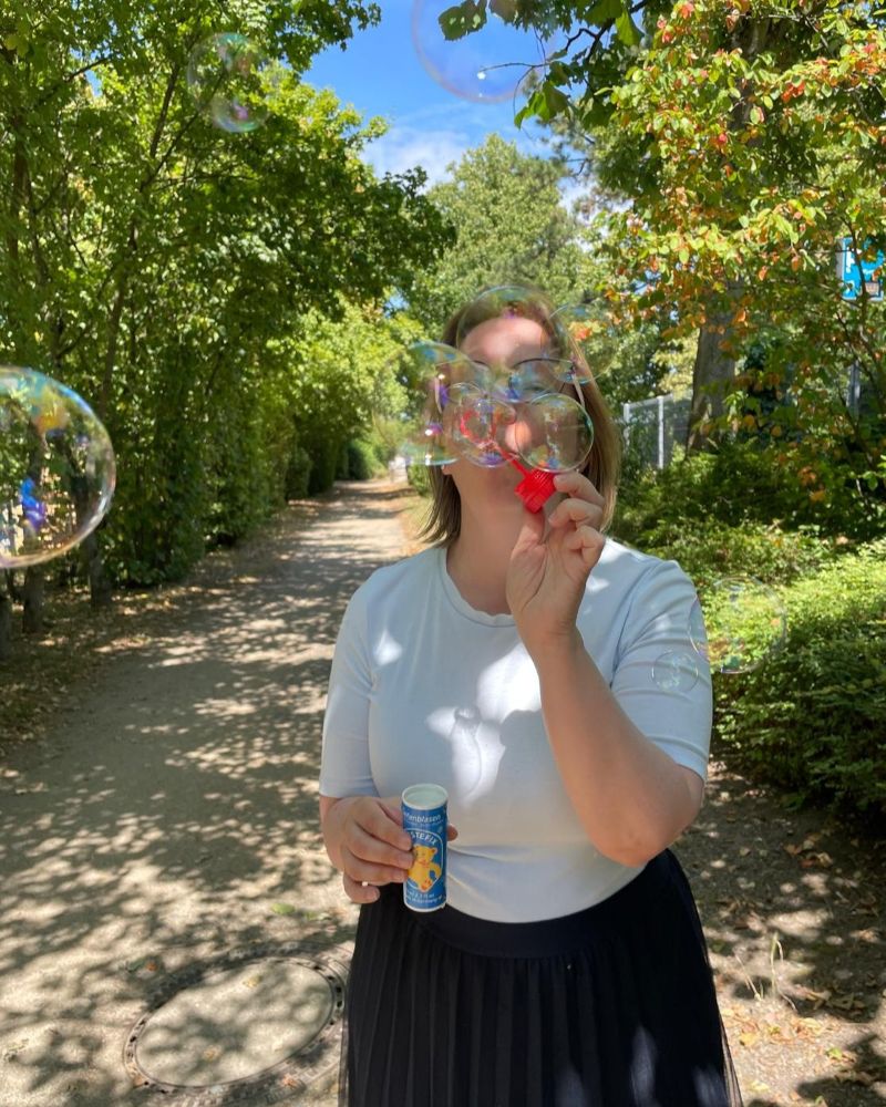 Astrid Steht auf einem lichtdurchfluteten Weg mit einer Flasche Pustefix. Vor Ihrem Gesicht und überall auf dem Foto sind Seifenblasen.