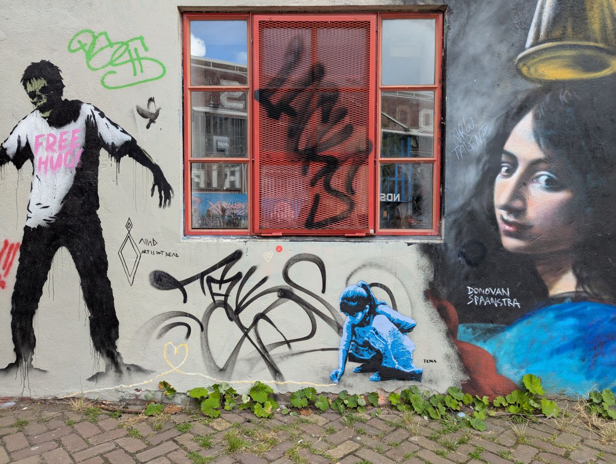 Grafittiwand auf dem NDS Areal in Amsterdam: Ein Zombie mit "Free Hugs"-Shirt, eine Friedenstaube, ein Bild von einer Frau mit dunklen Haaren in einem blauen Kleid und ein Kind, dass mit Kreide ein Herz malt.