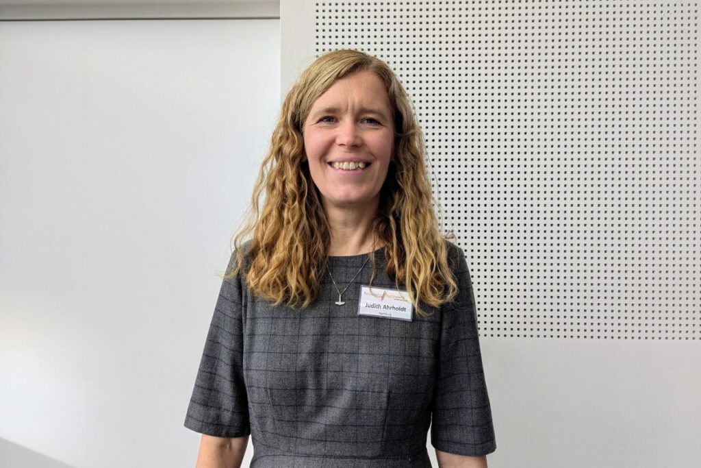 Judith Ahrhold lächelt entspannt in die Kamera, Sie trägt ein graues Kleid, die Haare sind offen und man sieht ihr Namensschild, denn das Foto ist am Rande der Assistants' World entstanden, wo sie den Stand des ANiD betreut hat.