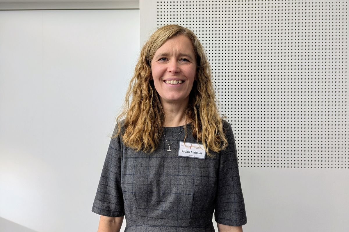 Judith Ahrholdt lächelt entspannt in die Kamera, Sie trägt ein graues Kleid, die Haare sind offen und man sieht ihr Namensschild, denn das Foto ist am Rande der Assistants' World entstanden, wo sie den Stand des ANiD betreut hat.