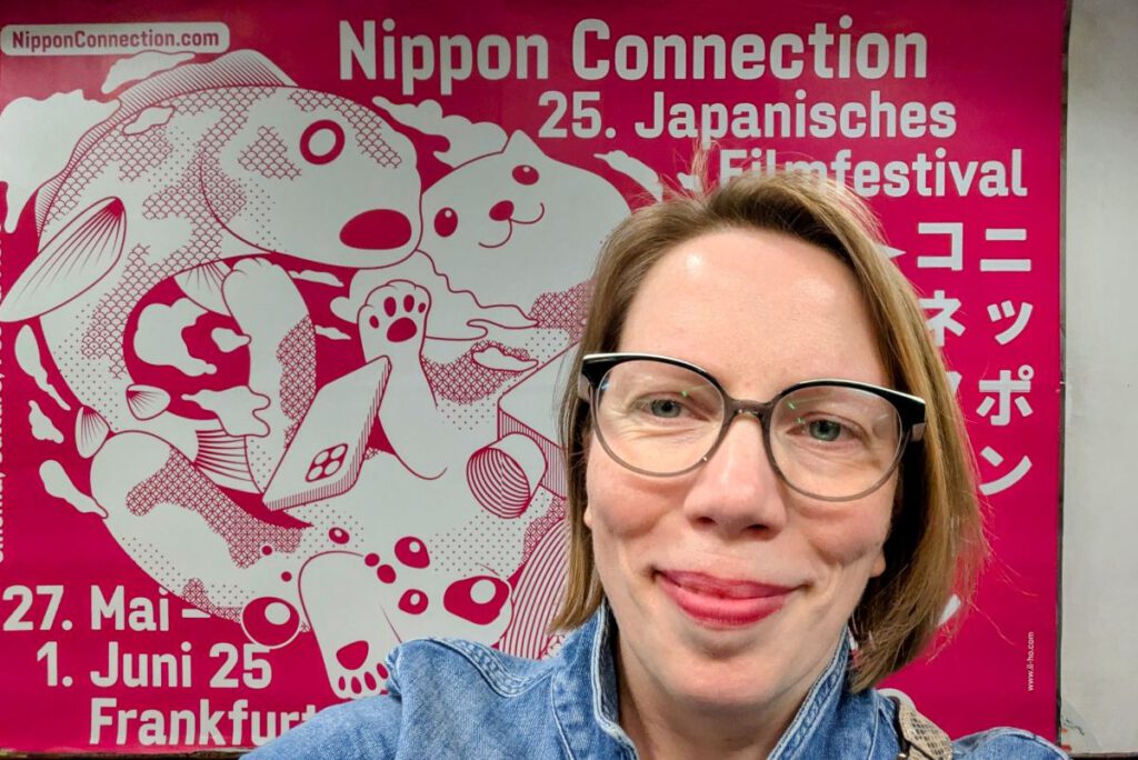 Astrid vor dem großen Nippon Connection Plakat in der B-Ebene des Willy-Brandt-Platzes. Lippenstift passt zum Plakat.