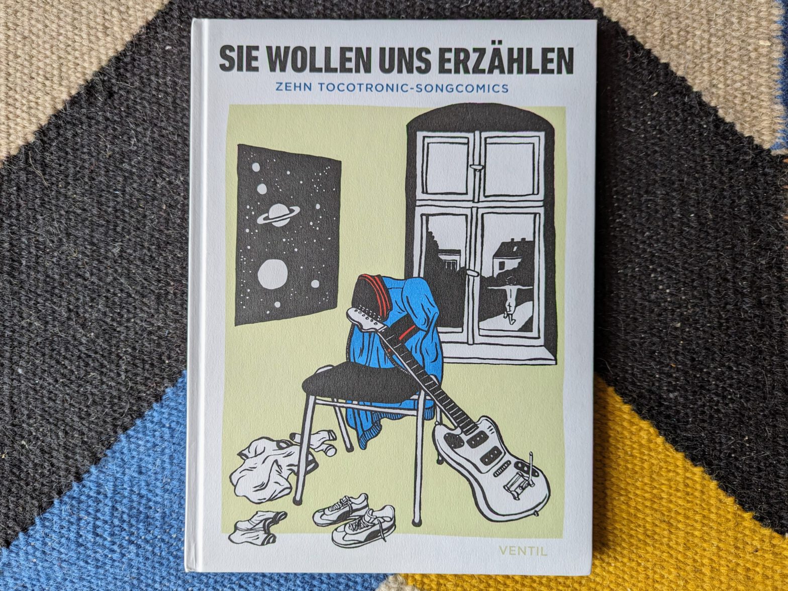 Foto des Buchcovers "Sie wollen uns erzählen. Zehn Tocotronic-Songcomics" auf einem bunten Teppich