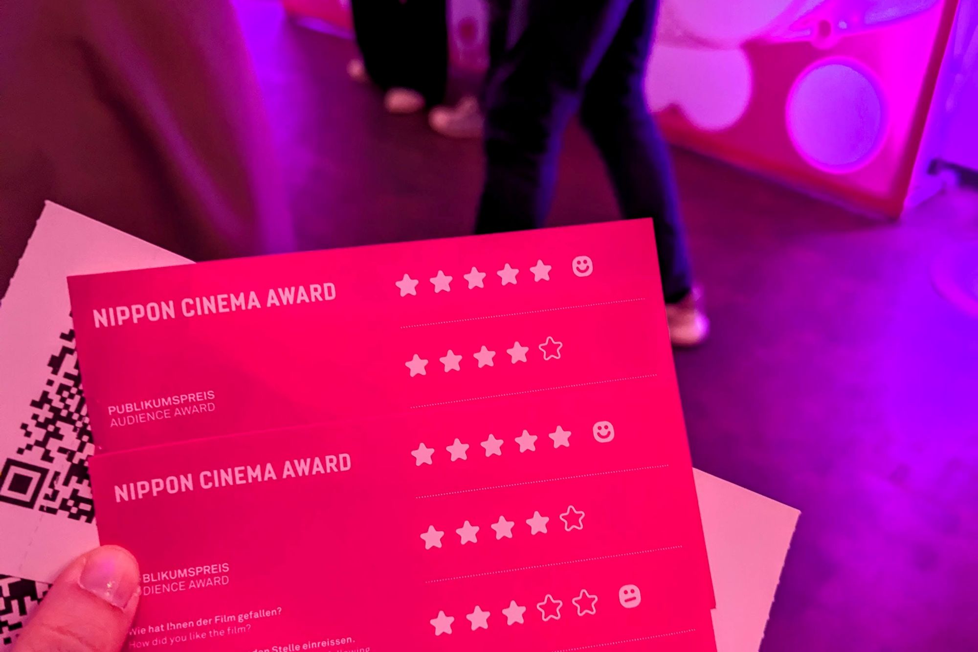 Ein Schnappschuss der Nippon Connection mit einer Hand, die zwei Kinokarten und zwei Stimmzettel für den Nippon Cinema Award in der Hand hält, alles ist in Festival-Pink gehalten.